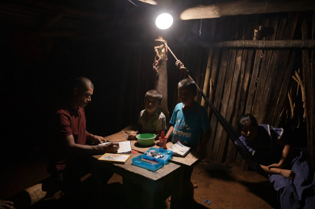 A father is helping his children (two boys and a girl on the side) with their homework at night, thanks to the use of the third generation solar home systems.